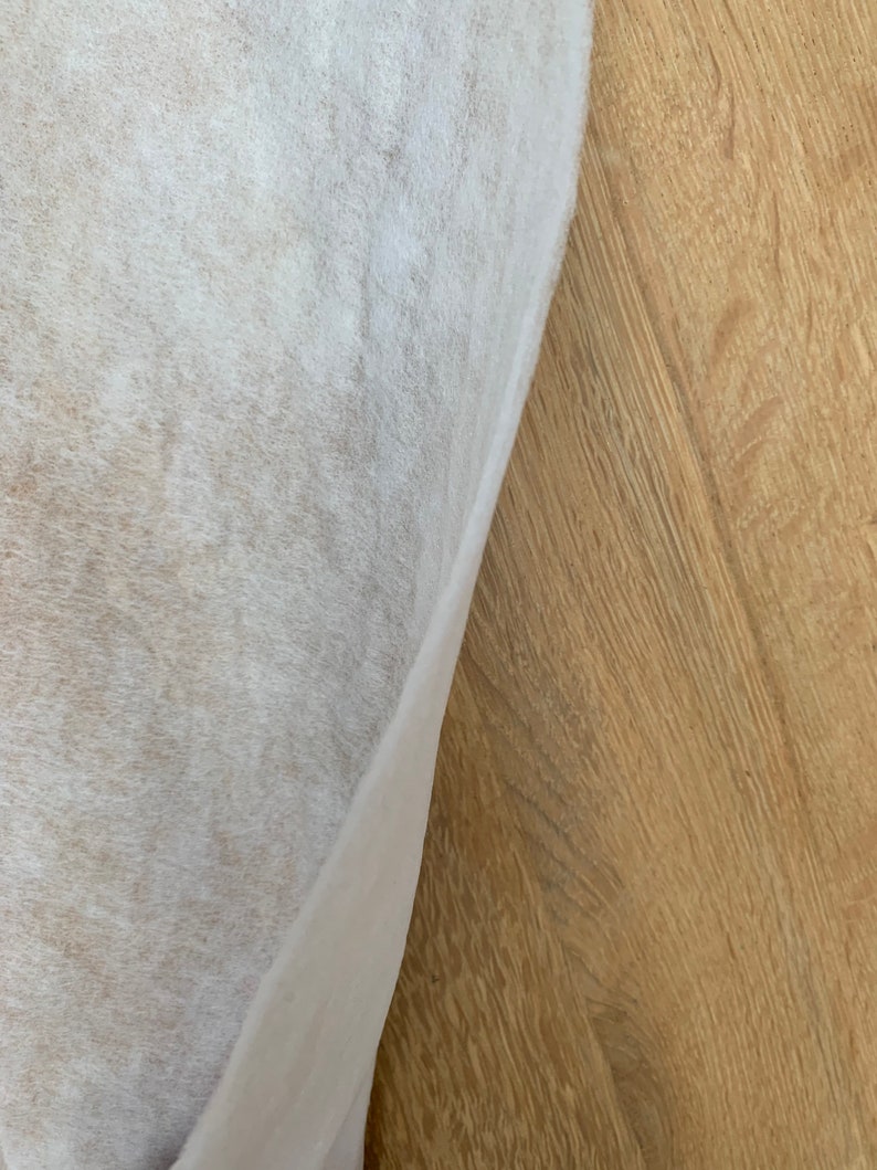 May include: A close-up view of a white, textured fabric partially covering a light brown wooden surface. The fabric has a soft, slightly translucent appearance, with visible fibers. The wood grain is prominent, creating a contrast in texture and color.