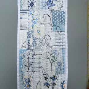 May include: A white fabric wall hanging with a patchwork design featuring blue and white floral patterns. The fabric is stitched together with white thread and the flowers are outlined in black thread.