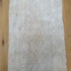 May include: A rectangular, off-white, non-woven fabric sheet. The sheet has a slightly textured surface and a light brown hue in the center. The fabric is placed on a wooden surface.