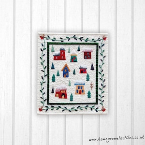 May include: A white quilted wall hanging with a green holly border. The centre of the quilt features a winter village scene with small houses, trees, and snow.