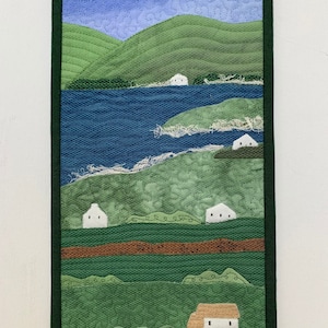 May include: A quilted wall hanging depicting a landscape with rolling green hills, a blue lake, and white houses. The houses are scattered throughout the landscape, with some nestled in the hills and others near the lake. The sky is a light blue with white clouds.