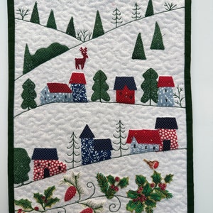 May include: A quilted wall hanging showing a winter village scene. The artwork features houses with red and blue roofs, evergreen trees, and a deer. The border is dark green, and the background is white with a quilted pattern.