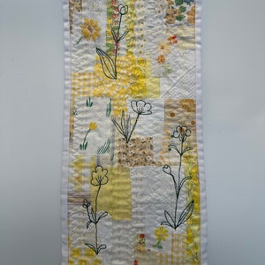 May include: A white fabric wall hanging with a patchwork design featuring yellow and white floral patterns. The fabric is stitched together in a grid pattern and has a white border. Black embroidery outlines simple flower designs on the fabric.