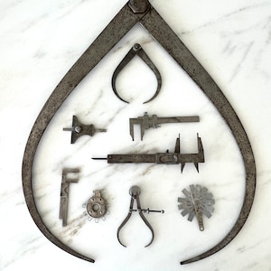 May include: An assortment of vintage metal measuring tools, including calipers, gauges, and a large divider, arranged on a white marble surface. The tools are various sizes and shapes, with a weathered, antique appearance.