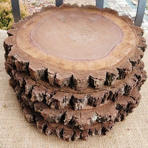 May include: A stack of four rustic, brown wooden coasters. Each coaster is a cross-section of a tree trunk, showcasing the natural grain and bark edges. The coasters are stacked on a burlap surface.