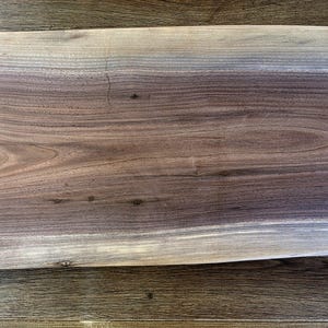 May include: A rectangular wooden cutting board with a natural edge. The board features a gradient of colors, from light tan to dark brown, showcasing the wood grain. The board is placed on a wooden surface.