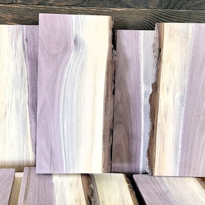 May include: A stack of five wooden boards with live edge, showing the natural grain and color variations of the wood. The boards are a mix of light and dark brown with purple hues.