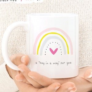 May include: White ceramic mug with a rainbow design in pink, yellow, and gray. The rainbow has a pink heart in the center and black dots. The text "a 'hug in a mug' for you" is printed below the rainbow.