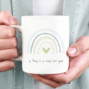 May include: White ceramic mug with a rainbow design in shades of blue and green. The mug has black dots and a green heart in the center of the rainbow. The text "a 'hug in a mug' for you" is printed on the mug.