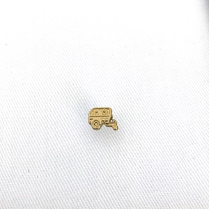 May include: A gold-coloured metal charm in the shape of a camper trailer. The charm is small and detailed, with a rounded roof and windows.