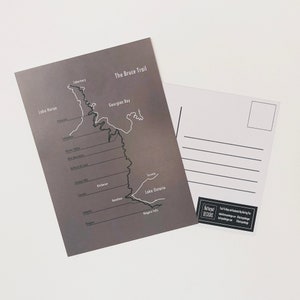 Könnte beinhalten: Eine graue Postkarte mit einer weißen Umrisskarte des Bruce Trails in Ontario, Kanada. Die Karte zeigt den Verlauf des Trails von Tobermory bis zu den Niagarafällen, vorbei an der Georgian Bay, dem Lake Huron und dem Lake Ontario. Der Text "The Bruce Trail" ist oben auf der Postkarte gedruckt.
