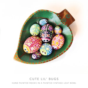 May include: A green leaf-shaped bowl with a brown rim, filled with hand-painted rocks shaped like ladybugs. The rocks are decorated with colorful dots and flowers. The bowl has a white background.