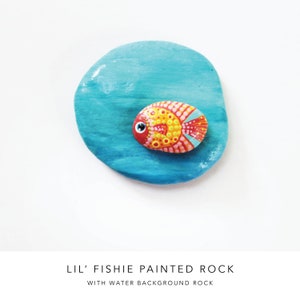 May include: A hand-painted rock depicting a small orange and red fish with white and yellow details. The fish is painted on a blue rock, resembling water.