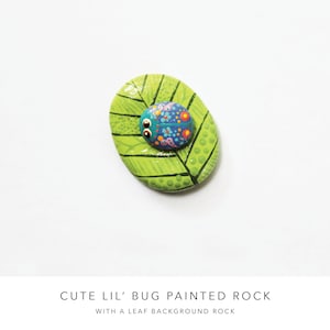 May include: Hand-painted rock with a green leaf design and a small blue, purple, and pink bug on top.