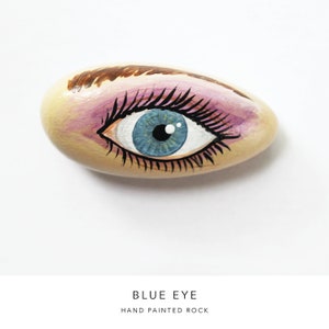 May include: Hand painted rock with a blue eye, pink eyeshadow, and brown eyebrow.