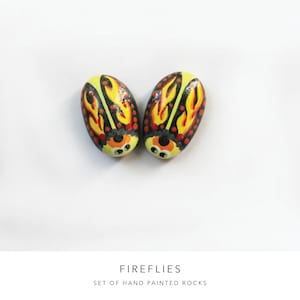 May include: Two hand-painted rocks shaped like fireflies with yellow, orange, black, and red details. The rocks are on a white background.  FIREFLIES SET OF HAND PAINTED ROCKS