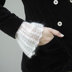 Pode incluir: Casaco de veludo preto com punhos de renda branca. Os punhos apresentam um delicado padrão vazado. O casaco tem botões brancos e um bolso. A estética geral é elegante e de inspiração vintage.