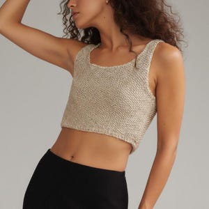 Könnte beinhalten: Ein beige gestricktes Crop-Top mit quadratischem Ausschnitt. Das Oberteil besteht aus einem strukturierten, gewebten Material. Das Model trägt einen schwarzen Unterteil. Das Oberteil ist ärmellos.