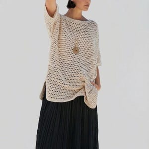 May include: A cream-colored, open-weave crochet top with short sleeves and a boat neck. The top has a relaxed fit and side slits. A wooden pendant necklace is visible. The top is paired with a black pleated skirt.