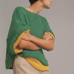 May include: A green knitted jumper with yellow edging on the sleeves and hem. The jumper has a loose, boxy fit and a round neckline. The sleeves are short and wide. The model is wearing beige trousers.