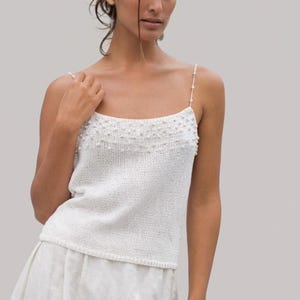 Könnte beinhalten: Weißes Strick-Camisole mit zarten Perlenriemen und einem perlenbesetzten Ausschnitt. Das ärmellose Oberteil ist perfekt für warmes Wetter. Das Camisole ist aus einem weichen, strukturierten Strickstoff gefertigt.