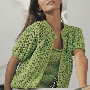 Może przedstawiać: Jasnozielony, ażurowy kardigan z krótkim rękawem i pasujący top. Kardigan ma zapięcie na jeden guzik. Strój wykonany jest z grubej przędzy, tworząc teksturowany wygląd. Ogólny styl jest swobodny i letni.