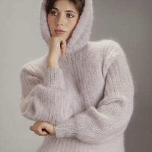 May include: A pale lilac hooded jumper with long sleeves. The jumper has a soft, textured appearance and a relaxed fit. The hood is up, and the sleeves have a slight balloon shape. The overall style is casual and comfortable.