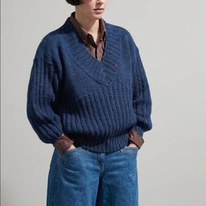 May include: A navy blue V-neck sweater with ribbed detailing and a small embroidered emblem. The sweater is paired with a patterned button-down shirt and blue jeans. The model is wearing a casual outfit.