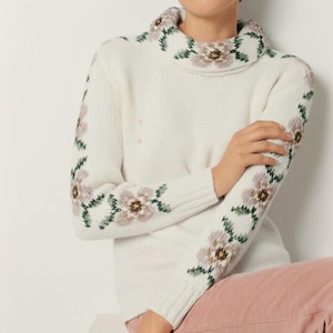 May include: Cream-colored turtleneck sweater with floral embroidery in shades of lavender, brown, and green. The floral pattern adorns the collar and sleeves. The sweater is made of a soft, knitted material.
