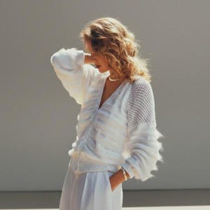 May include: A white knitted cardigan with a V-neck and button closure. The sleeves have a textured design and the right shoulder has a mesh pattern. The cardigan is paired with white trousers. A gold bracelet and a pearl necklace are visible.