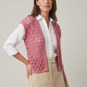 May include: A dusky pink crochet vest with an open weave pattern. The sleeveless top is worn over a white shirt and beige trousers. The vest's texture and colour create a soft, feminine appearance.