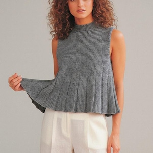 Könnte beinhalten: Ein ärmelloses, graues Strickoberteil mit hohem Ausschnitt und plissiertem Schößchen. Das Oberteil ist aus einem strukturierten Strickstoff gefertigt und wird mit einer weißen Hose kombiniert. Der Gesamtstil ist lässig und elegant.