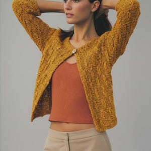 Op de afbeelding: Mosterdgele gebreide cardigan met een textuurpatroon en een enkele knoopsluiting. De cardigan wordt gedragen over een roestkleurige tanktop. Het model draagt ook een beige broek.