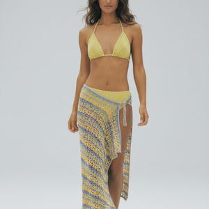 May include: A yellow crochet bikini top and a matching skirt with a high slit. The skirt features blue, yellow, and white stripes. The outfit is made of a lightweight, open-weave material, perfect for beachwear.
