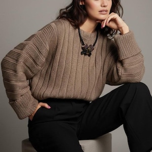 May include: A woman wearing a tan ribbed knit sweater with a round neckline and long sleeves. The sweater is paired with black pants. A black leather necklace with a decorative pendant is visible.