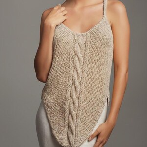 以下が含まれることがあります： Vネックと中央にケーブル編みのディテールが入ったベージュのニットタンクトップ。細いストラップと、わずかに非対称の裾があります。生地は柔らかい天然繊維のようです。