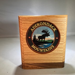 May include: A wooden plaque with a circular patch featuring a moose silhouette, mountains, and a lake. The patch is surrounded by a black circle with the text "ADIRONDACK MOUNTAINS" in gold lettering.