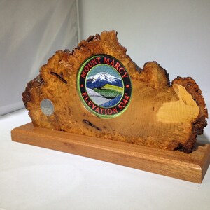 May include: A wooden plaque with a circular patch featuring the text "Mount Marcy Elevation 5344" and a depiction of a mountain with snow on the peak. The plaque is mounted on a wooden base.