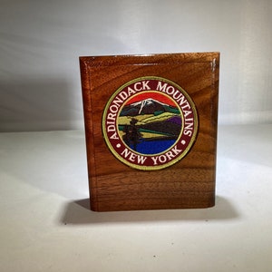 May include: A wooden plaque with a circular embroidered patch that reads "Adirondack Mountains New York". The patch features a mountain range with a red sunset and a blue lake.