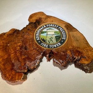 May include: A wooden slice with a round, black and white patch that reads "Adirondack Forest Preserve Fire Tower". The patch features a small, green and white illustration of a fire tower.