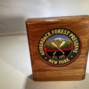 May include: A wooden box with a circular patch on the front. The patch is black with white text that reads "Adirondack Forest Preserve Est. 1885 New York". The patch also features a red canoe with two crossed oars and a green mountain in the background.