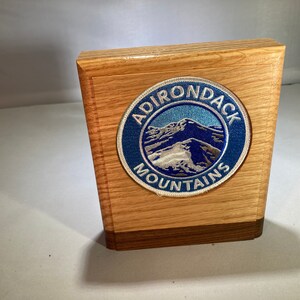 May include: A wooden box with a round patch featuring the words "ADIRONDACK MOUNTAINS" in white lettering. The patch has a blue border and depicts a mountain range in shades of blue and gray. The box is made of light brown wood.