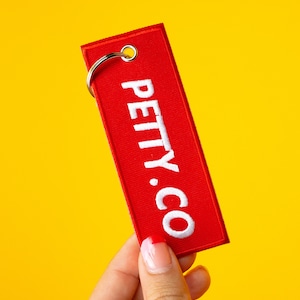 May include: Red fabric keychain with a silver ring and white embroidered text that reads "PETTY.CO"