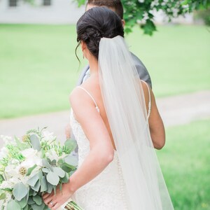 May include: A bride in a white lace wedding dress with a long veil walks down a path with her groom. She is carrying a bouquet of white flowers and greenery.
