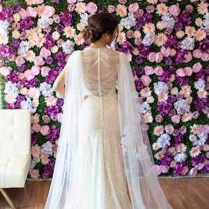 May include: A white wedding dress with a lace bodice and a tulle cape. The dress is shown from the back and the bride is standing in front of a wall of pink and purple flowers.