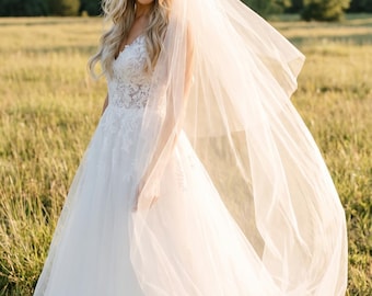 May include: A white wedding dress with a flowing tulle skirt and a detailed bodice. A sheer veil is draped over the shoulders. The bride is standing in a field of tall grass, with trees in the background.