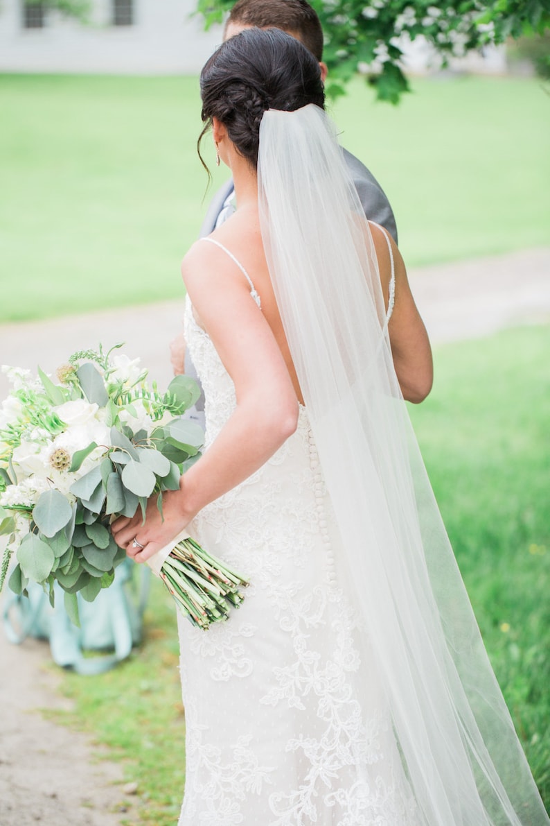May include: A bride in a white lace wedding dress with a long veil walks down a path with her groom. She is carrying a bouquet of white flowers and greenery.