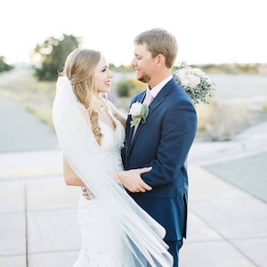 May include: A bride and groom stand close together, smiling and looking at each other. The bride is wearing a white wedding dress with a long veil. The groom is wearing a dark blue suit with a pink tie. The couple is standing outdoors in front of a blurred background.