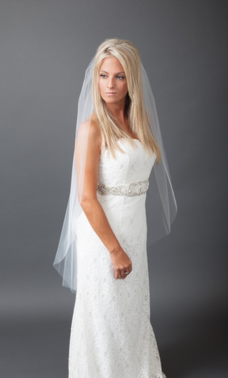 May include: A bride wearing a white lace wedding dress with a beaded belt and a white veil. She is standing against a gray background.