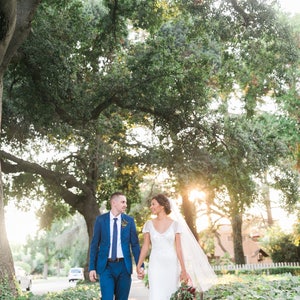 May include: A bride and groom walking hand-in-hand down a path lined with greenery. The bride is wearing a white lace dress with a veil and carrying a bouquet of red flowers. The groom is wearing a blue suit with a blue tie. The couple is smiling and looking at each other.